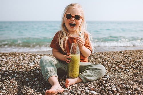 Snapshot of happy blonde girl in sunglasses, sitting on a beach and smiling while holding a tropical smoothie