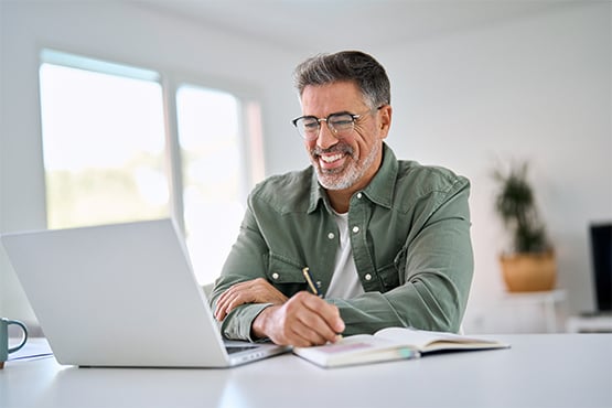 Smiling silver-haired man in glasses, checking his high interest checking account balance on his laptop at home in Fargo, ND