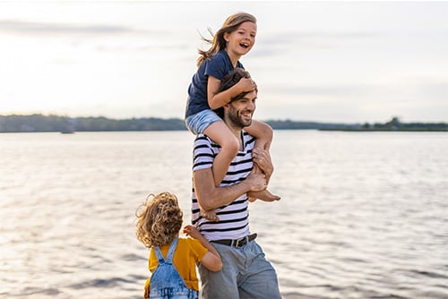 Young brunette girl laughing on her dad’s shoulders as they walk along the edge of the water in Devils Lake, ND