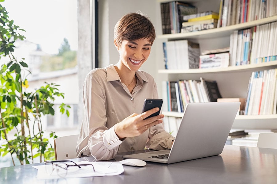 a happy businesswoman sits at her desk with a phone and laptop and enjoys the benefits of positive pay from Gate City Bank