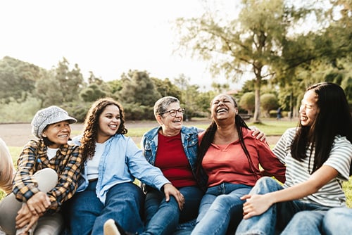 Five laughing women of different ages, sharing stories while sitting on the grass in jeans in Roosevelt Park in Minot, ND