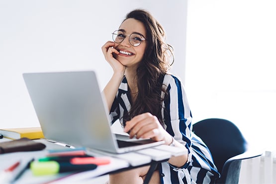 a happy woman wearing glasses leans on her hand and glances left while using her laptop to see Gate City Bank’s IRA options