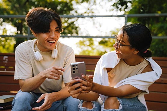 Joyful teen friends exchange funds over a cash app while sitting on a park bench in Grand Forks, ND.