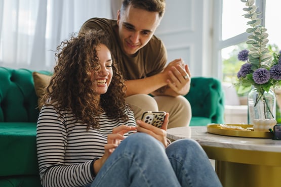 Curly-haired woman using her Gate City Bank mobile app beside her boyfriend on a green couch inside their North Dakota home