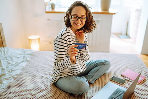 Curly-haired young woman, sitting on her bed and smiling while using her Gate City Bank debit card to shop online