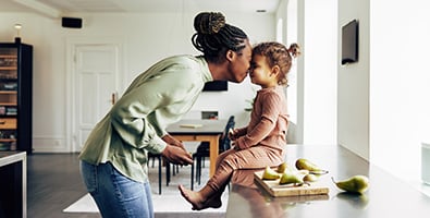 Loving mom touches noses with her preschool-age daughter, as she slices pears on the kitchen counter of their new home